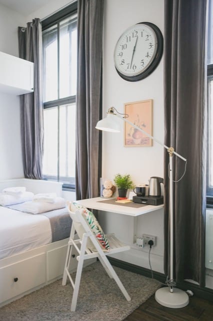 A pull out work desk that lays flat against the wall along with a foldable chair, next to a small double bed