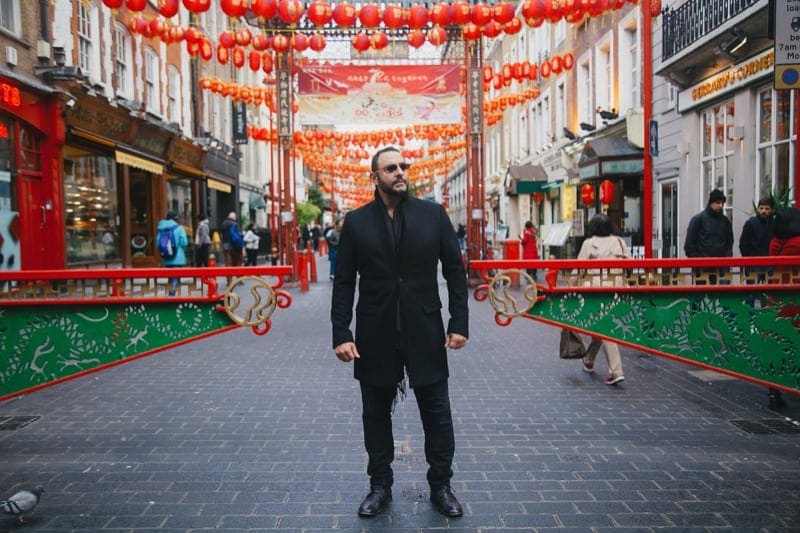 Man in a black overcoat and sunglasses stands in the middle of china town, london and poses for his holiday photos