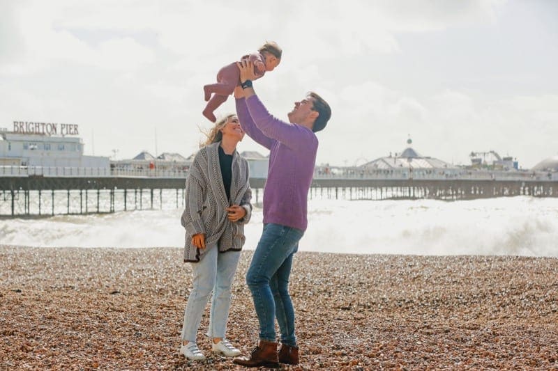 father lifts young baby in the air on brighton beach while mother watches smiling for their perfocal travel photographer