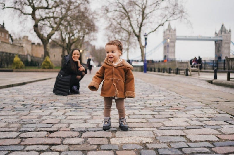 Tolder in brown jacket and tights standing on south bank london, whilst mum watches in the background for their holiday photoshoot