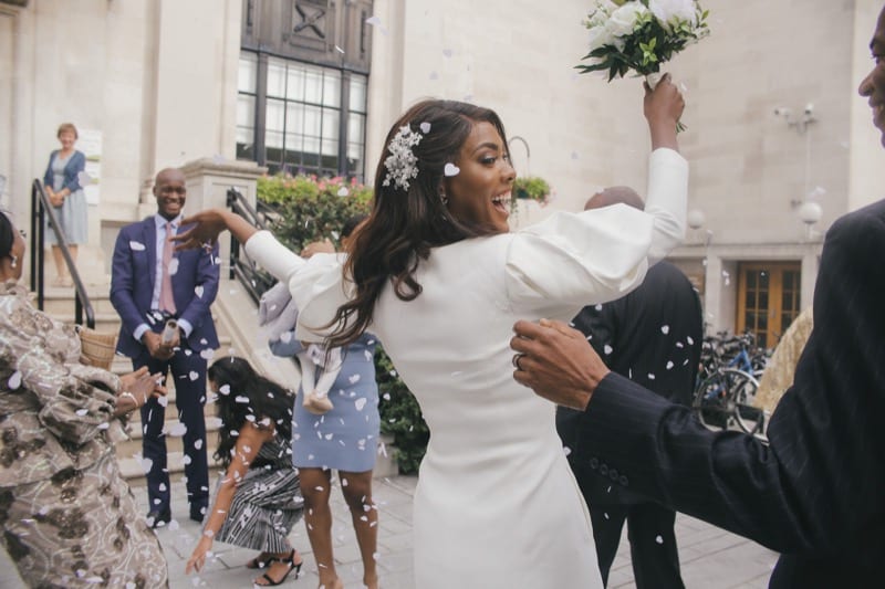 Black woman in white peticoat wedding dress looks over shoulder and throws bridal bouquet surrounded by confetti and family